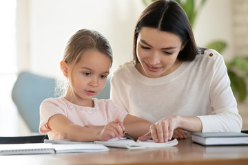 Fototapeta premium Caring young mom sit at table with small preschooler daughter help with studying, loving mother or nanny teach little girl child, reading together or learning, early development, education concept