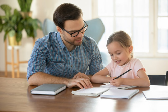 Caring young father and little preschooler daughter sit at table at home studying writing in notebook, loving dad or tutor teach small girl child handwriting, learning together, education concept