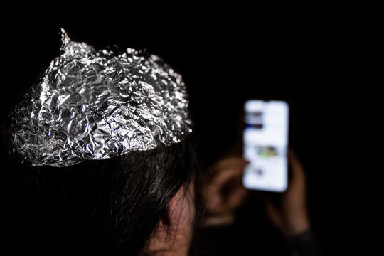 Man in the dark wearing a tin foil hat, smartphone in the hand, conspiracy theory and mind control