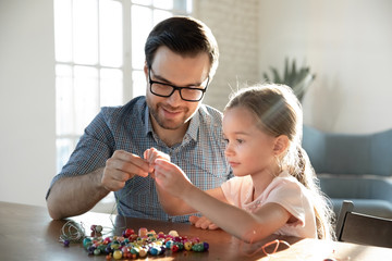Loving father sit at desk with little daughter have fun making bracelets with wooden beads together, caring dad and small preschooler girl child engaged in creative activity at home on family weekend