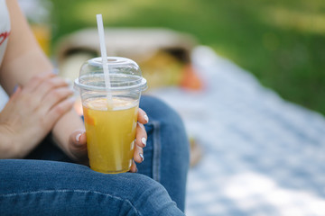 Close-up of woman hold disposable cup with orange lemonade on picnic outdoors. Space for text