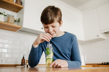 Teenager boy drinking infused detox water with cucumber, lemon and mint from glass bottle using...