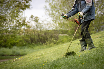 A man with a lawn mower mows grass and daisies on a slope near the house on a sunny spring day. A man in protective clothing, gloves and rubber boots mows the grass with a trimmer.