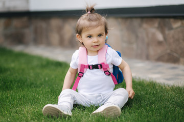 Adorable little girl sitting on the grass with backpack. Cute little girl outdoors