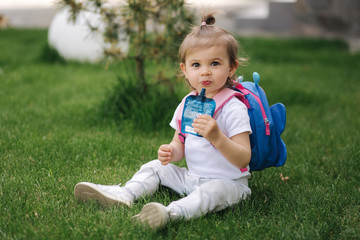 Adorable little girl sitting on the grass with backpack. Cute little girl drinking juise outdoors
