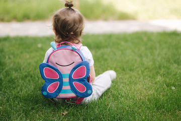 Back view of little girl with backpack sits on the grass outdoors. space for text