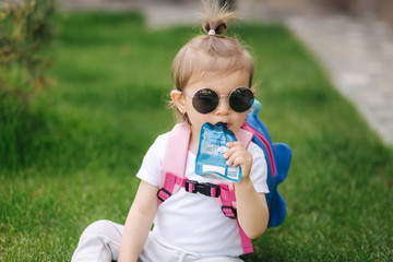 Adorable little girl sitting on the grass with backpack. Cute little girl drinking juise outdoors