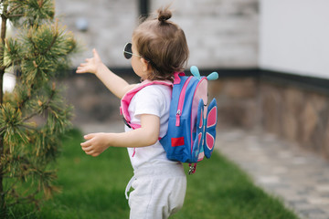 Portrait of Happy litle girl walk in gront of house with backpack. Cute one and half year girl in sunglasses