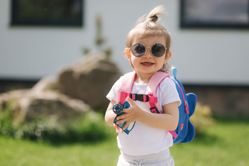 Happy litle girl walk in gront of house with backpack. Cute one and half year girl in sunglasses