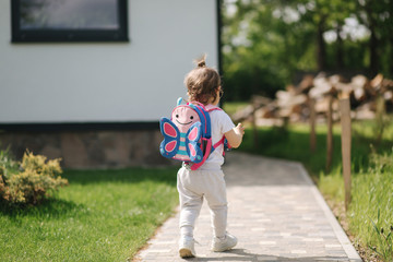 Happy litle girl walk in gront of house with backpack. Cute one and half year girl in sunglasses