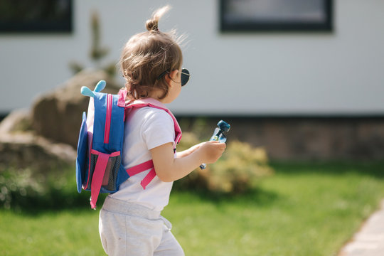 Happy litle girl walk in gront of house with backpack. Cute one and half year girl in sunglasses