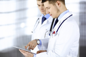 Two doctors with stethoscopes discuss medical exam resoults standing at hospital office. Physician and his colleague use a computer tablet for checking medication names records. Perfect medical