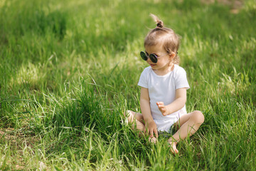 Happy little girl sits on the grass outdoors. Cute baby girl in white bodysuit and sunglasses on the backyard. Adorable kid play with grass