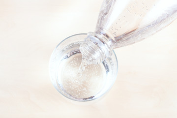 top view of pouring sparkling mineral water from transparent plastic bottle in glass on light brown...