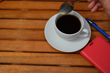a cup of black coffee in a white cup in the morning on wooden table with  red cover telephone and blue pen
