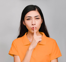 young beautiful asian woman,long black hair, wore orange t shirt, Pointing finger on mouth, Show The secret face,Do not disturb, on gray background