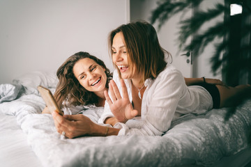Two young happy beautiful women romantic couple and lesbian family are lying in bed and chatting on a video chat in a smartphone, laughing merrily and enjoying life