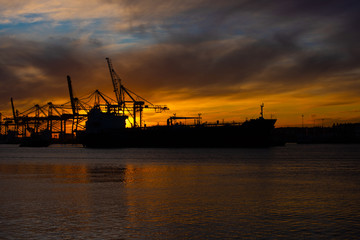 Silhouette of an oil tanker at sunset. Container port in the background