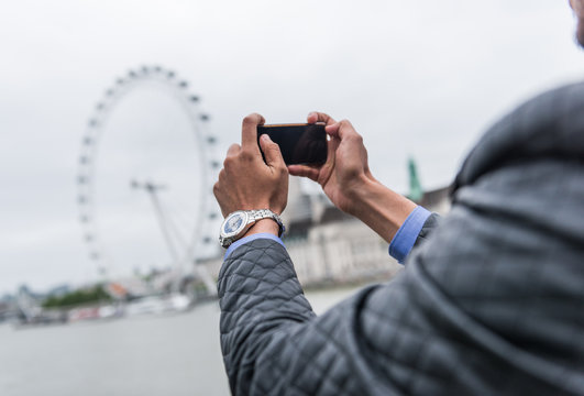 london, england, 05/05/2020 A male tourist from behind taking photos with his mobile phone outside a typical iconic london tourist attraction setting. The london eye is popular with tourists.