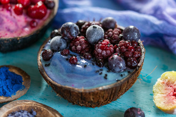 Coconut bowl of frozen raspberries and blackberries on a cyan worn out table top with blue cream. Smoothie bowl with frozen summer fruit.