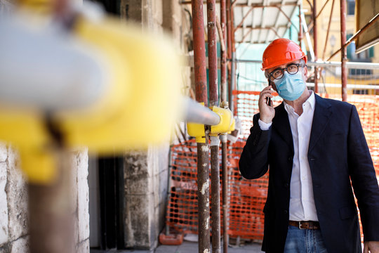 Ingegnere con casco arancione e mascherina Chirurgica lavora in un cantiere in citt&agrave; e parla con il cellulare 