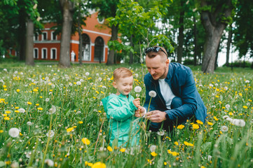 Cute happy little boy in green jacket takes white dandelion from father's hands.