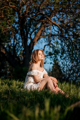 A beautiful young woman is sitting in a field in the green grass. She has a white dress, light hair, bare shoulders, and a happy face. Enjoy nature.