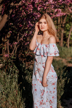 Beautiful girl with light hair in white clothes bare shoulders near blooming pink сercis flowers against the setting sun