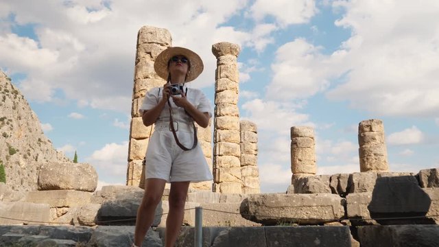 Young woman visiting ruins of temple of Apollo in Delphi, archaeological site by mount Parnassus, Greece, Europe. Large hat, fashion white dress.