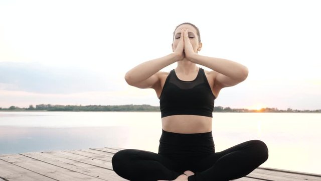 Adult woman do yoga meditation sitting in lotus position on boardwalk, deep enjoys physical, mental relax with closed eyes near water outdoors alone for calm mind. Practice asana exercise for wellness