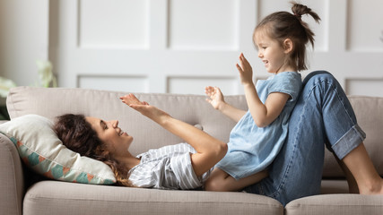 Smiling mother playing funny game with little daughter, clapping hands, lying on cozy couch at...