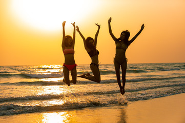 Silhouette group of happy young beautiful Asian women in bikini swimwear playing and jumping together on tropical beach at sunset. Three sexy girls friends having fun in summer holiday vacation travel