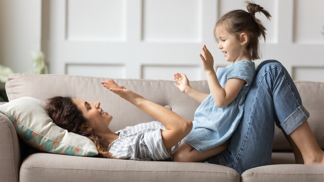 Smiling mother playing funny game with little daughter, clapping hands, lying on cozy couch at home, cute preschool girl having fun with loving young mum, family enjoying leisure time together