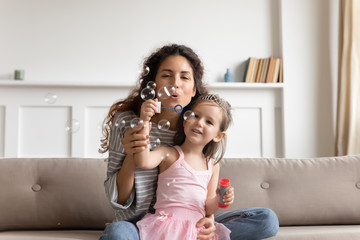 Happy mother and cute little daughter blowing soap bubbles, sitting on cozy couch in living room,...