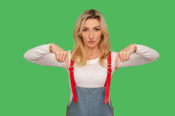 Here and right now! Portrait of angry tyrant bossy adult woman in denim overalls controlling and demanding to immediately approach her, pointing down. indoor studio shot isolated on green background