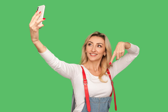 Portrait of happy adult woman with friendly smile chatting on video call and pointing finger down, meaning look below and subscribe while streaming on smartphone. indoor studio shot, green background - Powered by Adobe