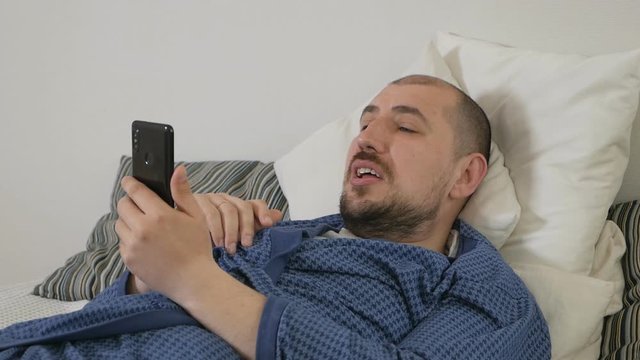 Lazy man using a mobile phone lying on a bed at home