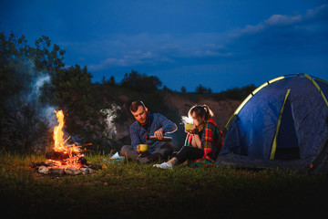 Night camping in the mountains. Happy couple travellers sitting together beside campfire and...