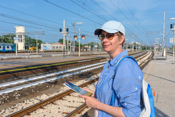 A middle-aged woman tourist with a backpack and a map in his hands at the railway station, against the background of rails. Travel around Europe by train.