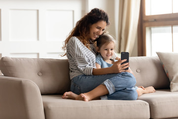 Smiling mother and little daughter hugging, using phone together, taking selfie, sitting on cozy couch in living room, happy young mum and preschool girl playing mobile game, making video call