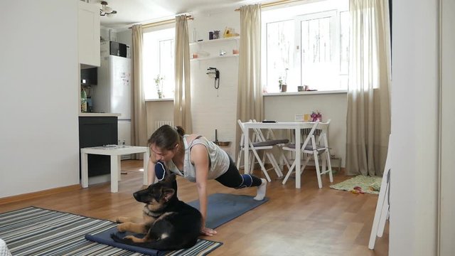 The girl does exercises while watching a program on TV at home. A young woman in sportswear, athletic build, plays sports in the room, in front of the TV. Her favorite pet, a dog lies next to her