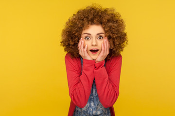 Oh my god, wow! Portrait of excited amazed woman with fluffy curly hair holding hands on face and looking with mouth open in surprise, shocked expression. indoor studio shot, yellow background