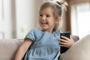 Close up smiling overjoyed funny little girl holding phone, having fun with mobile device, sitting on cozy couch alone at home, playing games, watching cartoons, using smartphone apps