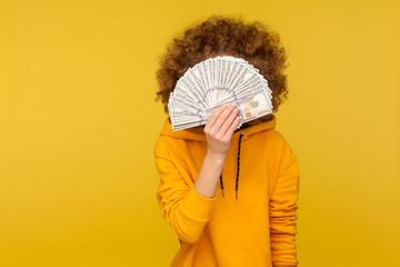 Curly-haired woman in urban style hoodie hiding face behind bunch of dollar banknotes, anonymous...