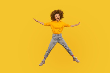 Portrait of enthusiastic excited curly-haired girl in urban style hoodie and jeans jumping high in air, flying and shouting with amazed happy expression, celebrating success. studio shot isolated