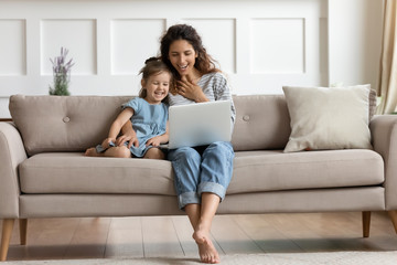 Happy mother and laughing little daughter looking at laptop screen, sitting on cozy couch, spending leisure time in living room, watching funny cartoons together, having fun with gadget at home