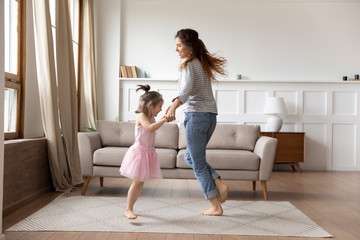 Happy mother and little daughter wearing princess dress holding hands, dancing in modern living...