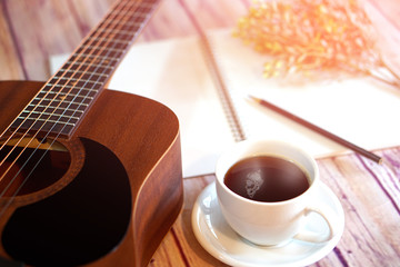 Acoustic guitar placed with a coffee cup, notebook and pencil placed on a wooden floor.