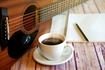 Acoustic guitar placed with a coffee cup, notebook and pencil placed on a wooden floor.