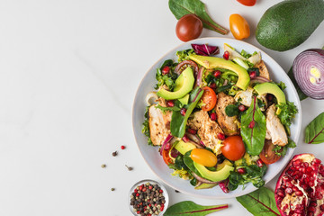 Healthy salad with grilled chicken breast, avocado, pomegranate seeds and tomato on white background. Top view